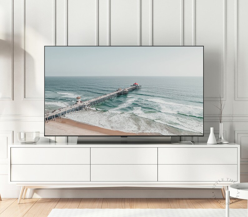 May include: A television displaying a photograph of a pier extending into the ocean with waves crashing against the shore. The pier is made of wood and has a red roof. The water is a light blue color and the sky is a light gray color.