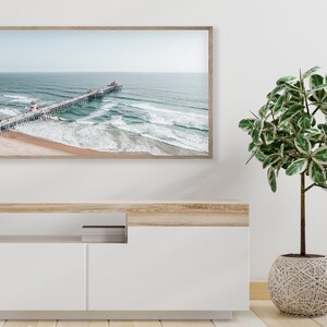 May include: An aerial view of a wooden pier extending out into the ocean with blue water and white waves crashing on the sandy beach. The pier has people walking on it and a red roof at the end.