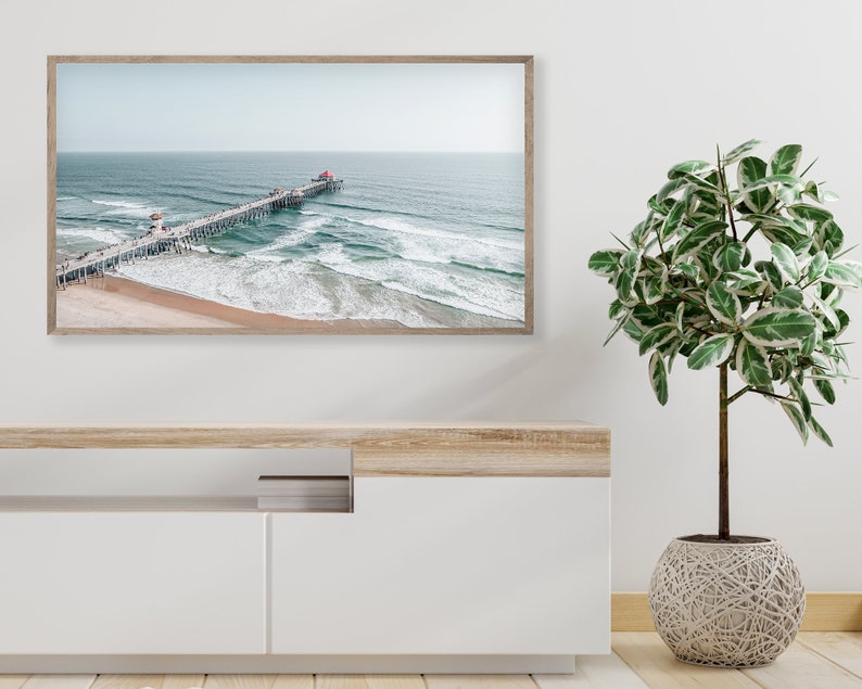May include: An aerial view of a wooden pier extending out into the ocean with blue water and white waves crashing on the sandy beach. The pier has people walking on it and a red roof at the end.