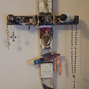 Könnte beinhalten: Ein Holzkreuz, das mit verschiedenen Gegenständen verziert ist, darunter eine kleine goldene Jesusfigur, ein Pinsel, eine Rosenkranz und ein kleiner Holzblock mit dem Buchstaben "O" darauf.