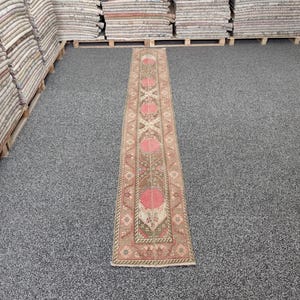 May include: A rectangular, patterned runner rug with a central design of repeating pink floral motifs and geometric shapes. The rug has a brown and beige background with red and green accents. The rug is displayed on a gray floor.
