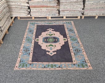 May include: A rectangular rug with a central geometric design in pink, green, and blue on a dark blue background. The rug has a light blue border with decorative elements and is displayed on a gray surface.