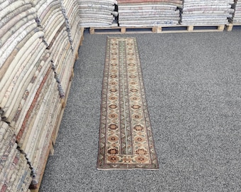 May include: A rectangular runner rug with a central floral pattern in beige, brown, and green. The rug has a border in a darker brown color. Stacks of rugs are visible on the left and in the background.