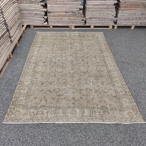 May include: A rectangular area rug featuring a muted beige and light brown color palette with a detailed floral and vine pattern throughout the center field and a decorative border. The rug has a low pile texture and short fringe on the ends.