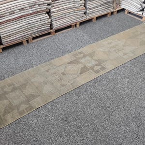 Peut inclure: Un long tapis de couloir étroit présentant une palette de couleurs beige et brun clair. Le tapis affiche un motif géométrique répétitif avec des formes en losange centrales et des motifs angulaires, au rendu délavé et usé.