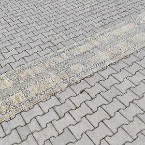 May include: A long, narrow, beige runner rug with a blue and white geometric pattern. The rug is laid out on a gray concrete surface.