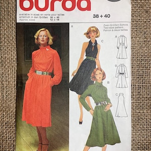 May include: Vintage Burda sewing pattern featuring illustrations of dresses. The cover displays a woman in an orange dress with a high neckline and a silver belt. The pattern includes sizes 38+40, with additional dress designs in black and green.