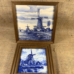 Può includere: Due piastrelle di Delft incorniciate, blu e bianche, che raffigurano mulini a vento in un paesaggio olandese. La piastrella superiore mostra tre mulini a vento con un canale in primo piano. La piastrella inferiore mostra un solo mulino a vento con un fiume in primo piano.
