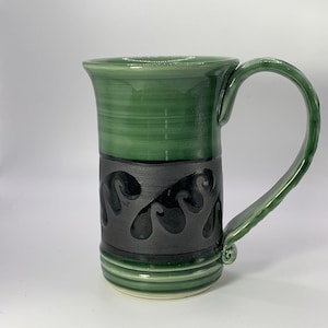 May include: A green ceramic mug with a black band and a swirling design. The mug has a handle and a rounded bottom.