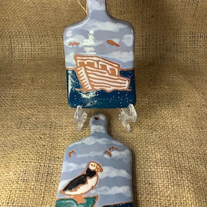 Peut inclure: Deux planches à découper en céramique sur le thème nautique. La planche du haut présente un bateau bleu et blanc avec une coque brune. La planche du bas présente un macareux bleu et blanc sur une falaise verte.