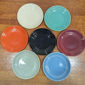 May include: A collection of seven small, round plates in various colors: cream, teal, orange, black, burgundy, light green, and blue. Each plate has concentric ring detailing and a smooth, glossy finish. The plates are arranged on a wooden surface.