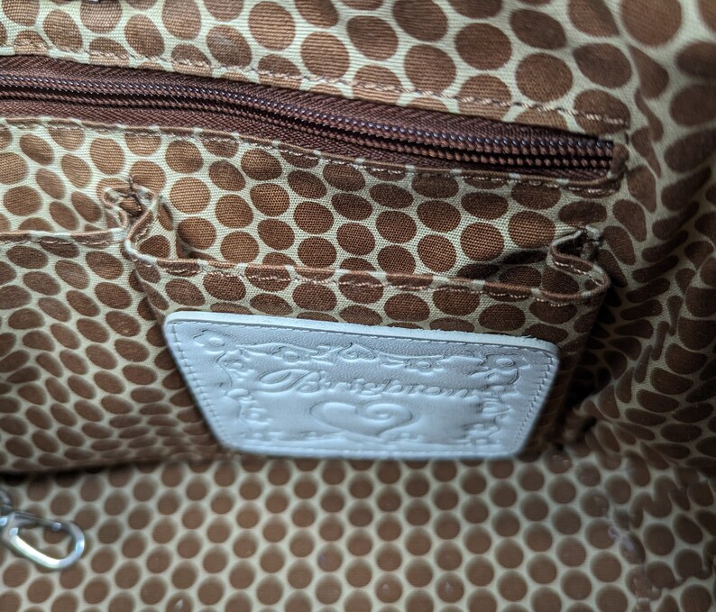 May include: Interior of a handbag with a brown polka dot lining and a zippered pocket. A white leather tag with the brand name "Brighton" is visible. The bag's interior also features small pockets.