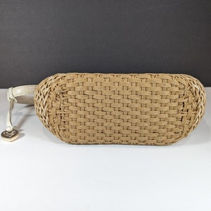May include: A woven beige handbag with a cream-colored strap and a gold heart-shaped charm. The bag has a rectangular shape with rounded edges and a woven pattern. The strap is attached to the side of the bag.
