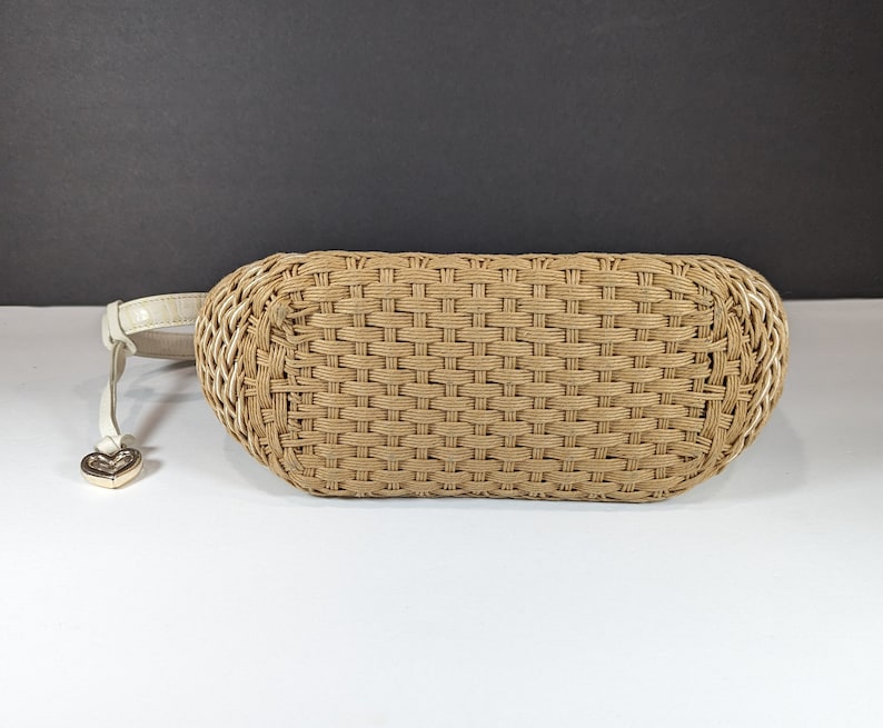 May include: A woven beige handbag with a cream-colored strap and a gold heart-shaped charm. The bag has a rectangular shape with rounded edges and a woven pattern. The strap is attached to the side of the bag.