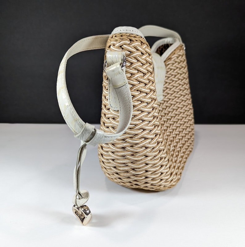 May include: Woven handbag with a cream-colored leather strap and a silver heart-shaped charm. The bag features a beige and white woven pattern. The strap is attached to the bag with silver hardware. The bag is set against a black and white background.