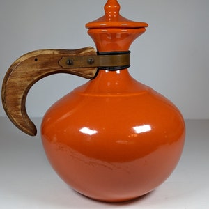 May include: Orange ceramic pitcher with a wooden handle and a brass band around the neck. The pitcher has a lid with a pointed top.