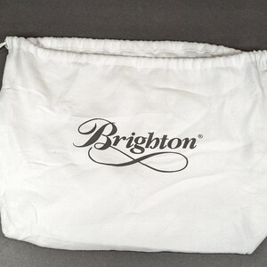 May include: A white drawstring bag with the Brighton logo in black script. The bag is made of a soft, textured fabric and has a drawstring closure at the top. The bag is empty and sits on a gray surface.