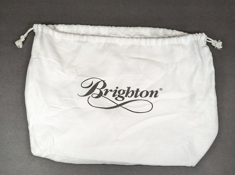 May include: A white drawstring bag with the Brighton logo in black script. The bag is made of a soft, textured fabric and has a drawstring closure at the top. The bag is empty and sits on a gray surface.
