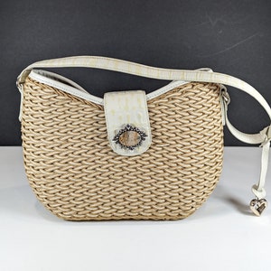 May include: Woven beige handbag with a white leather strap and accents. The bag features a decorative clasp with a stone-like center and a silver heart charm. The bag's body has a basket-weave design.