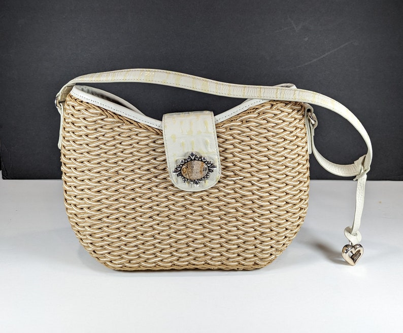 May include: Woven beige handbag with a white leather strap and accents. The bag features a decorative clasp with a stone-like center and a silver heart charm. The bag's body has a basket-weave design.