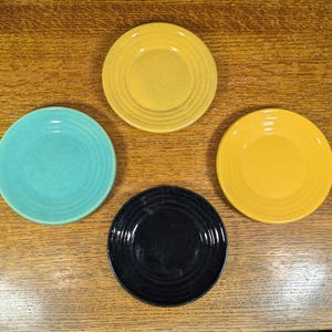 May include: Four small, round plates in shades of yellow, turquoise, and black are arranged on a wooden surface. Each plate has a slightly recessed center and concentric ring detailing. The plates offer a pop of color.