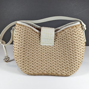 May include: Woven beige and white shoulder bag with a white leather strap. The bag features a textured weave pattern and a white flap with a crocodile skin texture. A silver heart-shaped charm hangs from the strap.