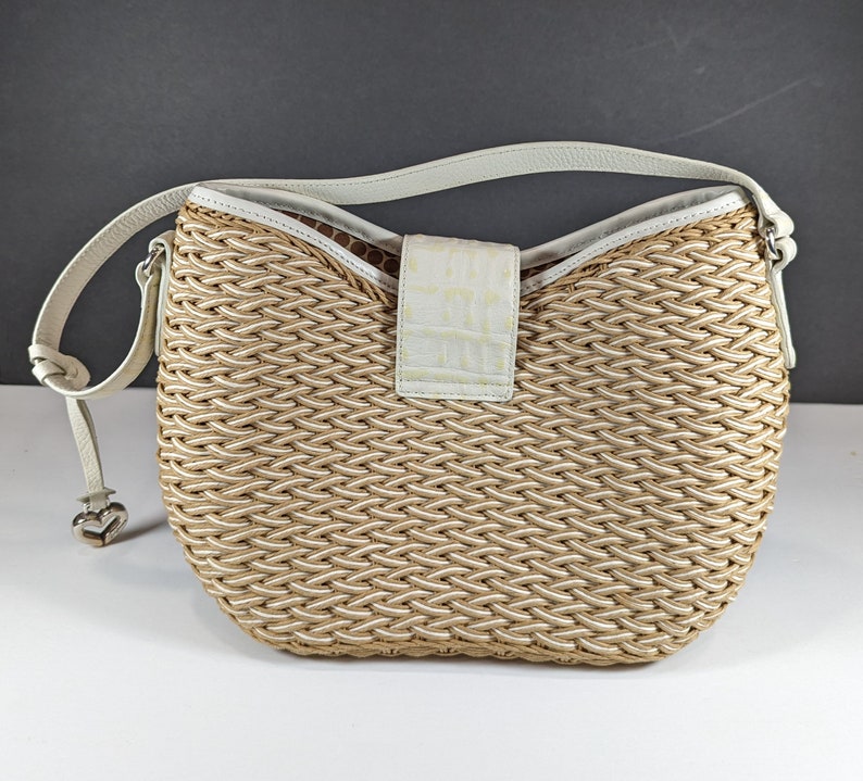 May include: Woven beige and white shoulder bag with a white leather strap. The bag features a textured weave pattern and a white flap with a crocodile skin texture. A silver heart-shaped charm hangs from the strap.