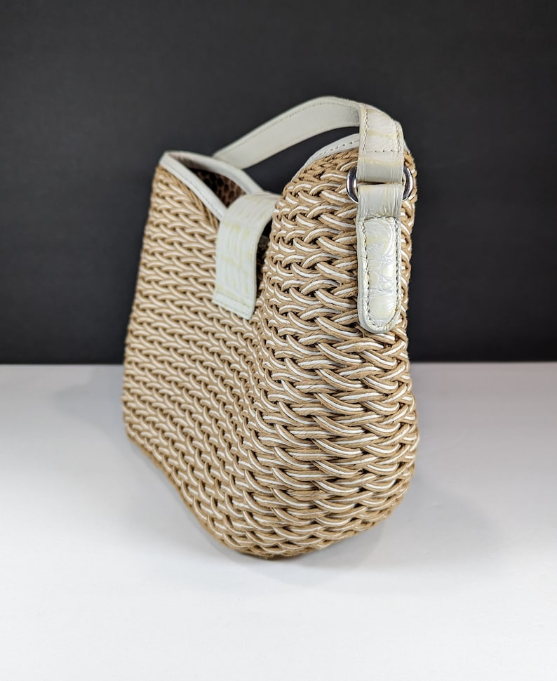 May include: A woven handbag with a beige and white pattern. The bag has a rounded shape and a white leather handle and trim. The bag is made of a thick, textured material and has a clean, modern design.