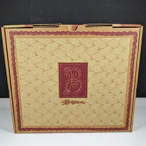 May include: A cardboard box with a woven pattern and a burgundy border. The center features a burgundy square with a stylized "B" and the brand name "Brighton" in a script font. The box is closed and appears new.
