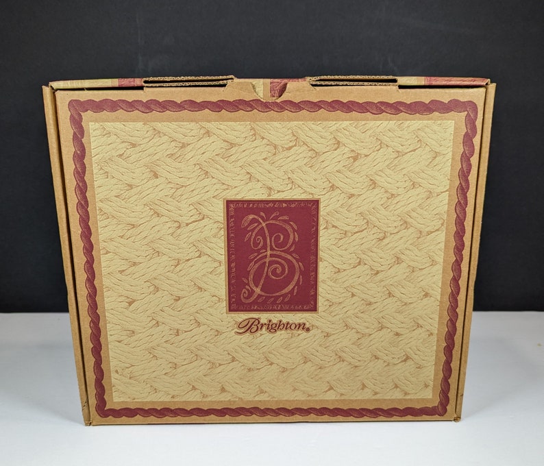 May include: A cardboard box with a woven pattern and a burgundy border. The center features a burgundy square with a stylized "B" and the brand name "Brighton" in a script font. The box is closed and appears new.