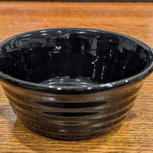 May include: A small, black ceramic bowl with a glossy finish. The bowl has a rounded shape with a ribbed exterior and two small handles. It sits on a wooden surface.