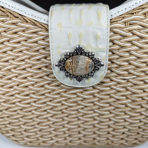 May include: Close-up of a woven handbag with a white leather strap and clasp. The clasp features a decorative silver-toned metal embellishment with a central stone. The bag's woven material is a mix of beige and white.