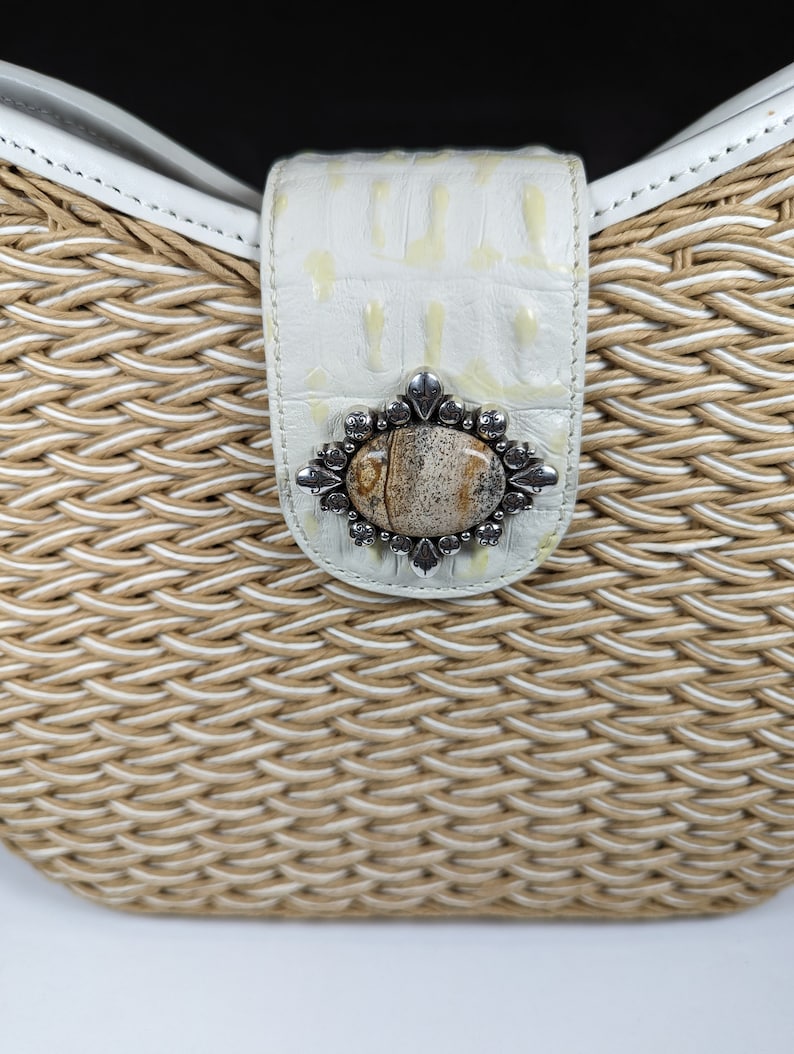 May include: Close-up of a woven handbag with a white leather strap and clasp. The clasp features a decorative silver-toned metal embellishment with a central stone. The bag's woven material is a mix of beige and white.