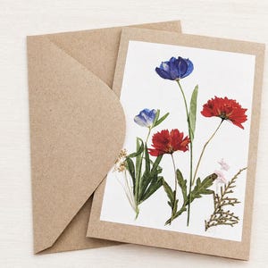 May include: A greeting card featuring pressed red and blue flowers with green stems and leaves, set against a white background. The card is inside a brown envelope, creating a natural and artistic aesthetic.