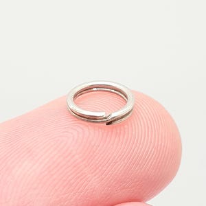 May include: A small, silver split ring, used for attaching items such as keys or charms. The ring is made of metal and has a split design for easy attachment. The ring is shown on a pink finger.