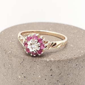 May include: A gold ring with a central clear gemstone surrounded by a cluster of small, round, deep pink gemstones. The ring's band is gold with etched geometric designs. The ring is set on a grey, cylindrical surface.
