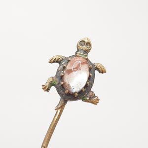 May include: A decorative hair pin featuring a turtle design. The turtle has a bronze-colored body with green accents and a central oval-shaped stone with red and white patterns. The pin's stem is a golden color.