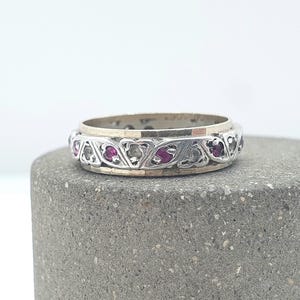 Könnte beinhalten: Ein silberner Ring mit einem dekorativen Band mit abwechselnd roten Edelsteinen und klaren Steinen. Der Ring liegt auf einer grauen, strukturierten Oberfläche und zeigt sein aufwendiges Design und seine Handwerkskunst.
