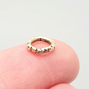 Op de afbeelding: Een kleine, cirkelvormige, zilverkleurige ring met een reeks gesneden, zich herhalende patronen rondom de omtrek. De ring ligt op een roze oppervlak, de achtergrond is een zachte, neutrale kleur.