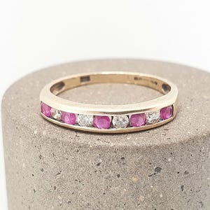May include: A gold ring featuring a series of alternating pink and clear gemstones. The ring is set on a grey, textured surface. The design is classic, suitable for everyday wear or special occasions. The ring's gemstones are round-cut.