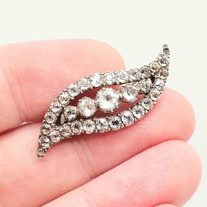 May include: A vintage-style brooch in a leaf shape, encrusted with clear, sparkling rhinestones. The brooch features three larger stones in the center, surrounded by smaller stones. The metal setting is silver-toned, creating a classic and elegant accessory.