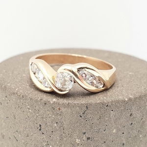 May include: A gold ring with a swirling design, featuring a large, round, clear gemstone at the center and smaller gemstones on either side. The ring is set on a gray, textured surface.