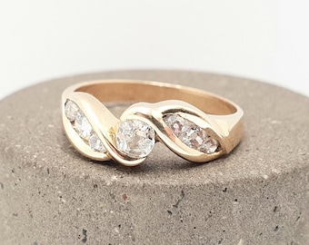 Könnte beinhalten: Ein goldener Ring mit einem wirbelnden Design, der einen großen, runden, klaren Edelstein in der Mitte und kleinere Edelsteine auf beiden Seiten aufweist. Der Ring liegt auf einer grauen, strukturierten Oberfläche.