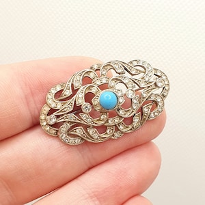 May include: A silver brooch with a turquoise stone in the center, surrounded by a delicate, intricate design of smaller stones.