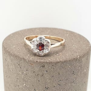 May include: A gold ring featuring a central round dark red gemstone surrounded by six clear white stones arranged in a floral cluster pattern. The band is thin and polished, with small etched line details on the shoulders near the setting.