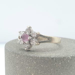 May include: A silver ring featuring a flower-shaped design. The center stone is a round, pink gemstone, surrounded by six clear, round gemstones. The ring is set on a gray, textured surface.