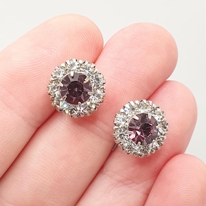May include: A pair of vintage stud earrings. Each earring features a central, round, dark purple stone surrounded by a halo of smaller, clear crystals. The earrings are set in a silver-toned metal and have a classic, elegant design.