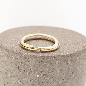 May include: A two-toned ring featuring a thin band with a metallic gold finish on one side and a silver-toned finish on the other. The band has an open, overlapping design where the two ends meet, creating a continuous circular shape with a textured surface.