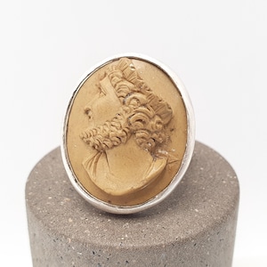 Op de afbeelding: Een zilveren ring met een camee die het profiel van een persoon met krullend haar afbeeldt. De camee is lichtbruin en gezet in een zilveren band. De ring wordt getoond op een grijs, cilindrisch oppervlak.