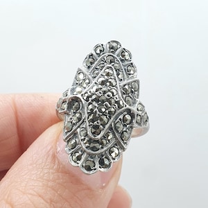 Op de afbeelding: Een zilverkleurige ring met een uitgebreid ontwerp. De ring heeft een centrale cluster van kleine, donkere stenen, omgeven door een gedetailleerd, symmetrisch patroon. De ring wordt tegen een effen witte achtergrond gehouden.